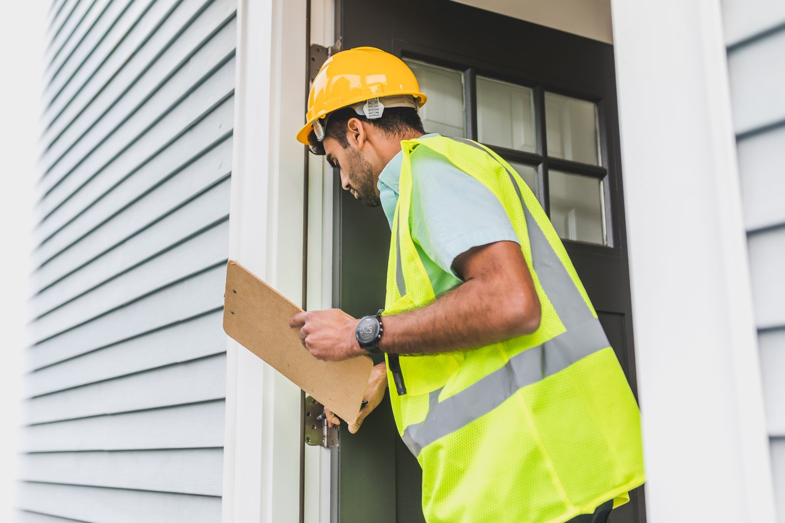 Home warranty inspection: inspector in a hard hat and yellow vest checking a house entrance with a checklist
